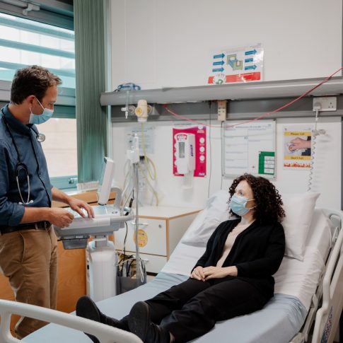 A woman with brown hair wearing a surgical mask lying on a hospital bed while a doctor with a surgical mask stands facing her